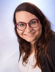 Frau Müller mit dunklem, langen Haar, Brille und hellem Lächeln vor blauem Verlaufshintergrund.